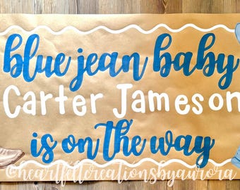以下が含まれることがあります： 茶色のバナーに青い文字で「Blue Jean Baby Carter Jameson is on the way」と書かれています。カウボーイハット、オーバーオール、カウボーイブーツのイラストが描かれています。