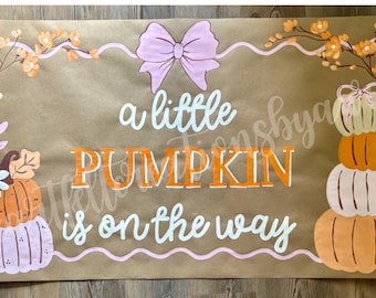May include: A brown banner with the text "a little PUMPKIN is on the way" in white and orange. The banner is decorated with painted pumpkins, flowers, and a bow, ideal for a baby shower or autumn decor.