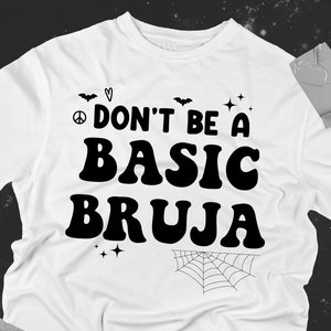 Könnte beinhalten: Weißes T-Shirt mit schwarzem Text, der "Don't be a basic bruja" lautet, mit einem Peace-Zeichen, Herzen, Fledermäusen und einer Spinnennetzgrafik.