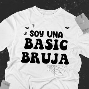 Könnte beinhalten: Weißes T-Shirt mit schwarzem Text, der "Soy una basic bruja" lautet, mit einem Peace-Zeichen, einem Herzen und einer Spinnennetzgrafik.