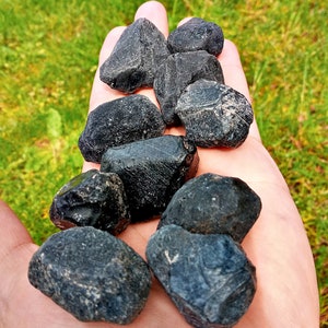 May include: A handful of rough, black tourmaline stones. The stones have a natural, unpolished appearance with varying shapes and sizes. The dark color contrasts with the green grass in the background. These stones are often used for their metaphysical properties.