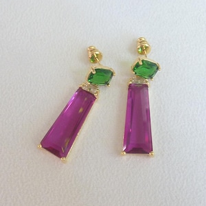 May include: A pair of gold-coloured earrings with magenta rectangular pendants. Each earring has a green, emerald-cut gemstone at the top. The earrings are displayed against a white background.