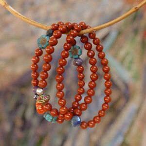 Op de afbeelding: Drie rode kralenarmbanden met decoratieve accenten, waaronder turquoise en paarse bloemen, en een gouden bedel. De armbanden hangen aan een houten tak.