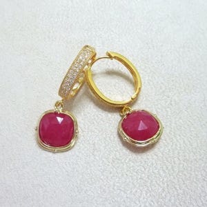 May include: A pair of gold-coloured hoop earrings featuring a square, faceted, ruby-red gemstone pendant. The hoop section is encrusted with small, clear crystals. The earrings are shown on a white surface.