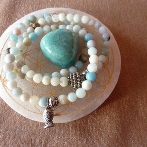 Puede incluir: Una colección de collares de cuentas y una piedra turquesa pulida sobre un plato blanco circular. Los collares presentan cuentas de color azul claro, crema y melocotón. Un dije de pez plateado cuelga de un collar. El plato tiene un patrón floral dorado.