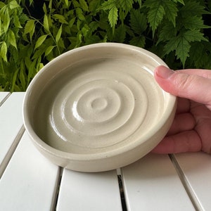 以下が含まれることがあります： 波打つ円形のデザインが施された白い陶器の皿。皿は少し傾いており、手作りのようです。