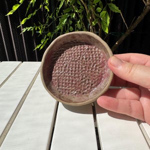 May include: A small, handmade ceramic soap dish with a textured, grid-like surface in shades of mauve and brown. The dish has a rounded shape and a raised rim. The soap dish is designed to allow water to drain, extending the life of the soap.