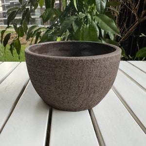 Pode incluir: Um vaso de cerâmica marrom escuro com uma superfície áspera e texturizada. O vaso é redondo e tem uma abertura larga.