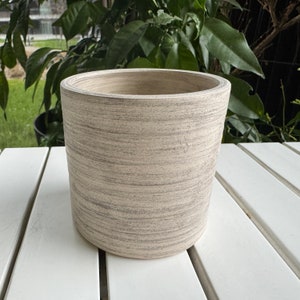 May include: A white ceramic planter with a textured, wood-grain finish. The planter is round and has a wide opening.