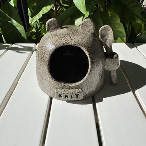 May include: A ceramic salt cellar shaped like a bear with a small spoon attached. The cellar is grey with black speckles and the word "SALT" is written on the front.
