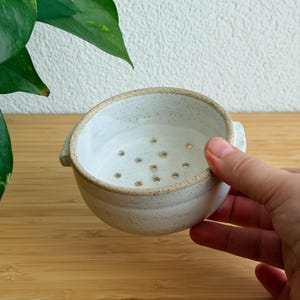 Könnte beinhalten: Eine kleine, runde Beeren-Schale aus weißer Keramik mit gesprenkelter Oberfläche und mehreren Abflusslöchern am Boden. Die Schale verfügt über zwei kleine seitliche Griffe und einen dicken, strukturierten Rand in handgefertigter Optik.