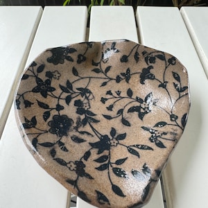 May include: A ceramic serving dish with a brown glaze and a black floral pattern. The dish has an irregular shape and a slightly raised rim.