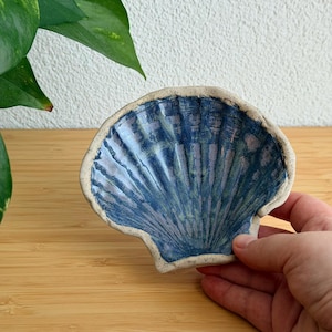 May include: A ceramic dish shaped like a scallop shell with a glossy blue and purple glaze. The edges of the shell are textured with a speckled off-white finish, featuring deep radial grooves that extend from the centre to the outer rim.