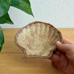Może przedstawiać: Ceramiczna miseczka w kształcie muszli przegrzebka z teksturowanym, nakrapianym szkliwem w kolorze kremowym i brązowym. Krawędzie muszli są podkreślone ciemnobrązową obwódką, a wewnętrzna powierzchnia posiada promieniste żłobienia imitujące muszlę.