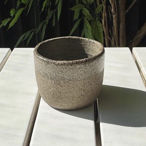 Könnte beinhalten: Eine handgefertigte, beige Keramiktasse mit strukturierter Oberfläche. Die Tasse hat eine abgerundete Form und einen leicht ausgestellten Rand. Der Innenraum ist dunkel, und der Rand hat einen dunkleren braunen Akzent. Die Tasse steht auf einer weißen Oberfläche.