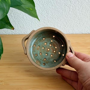 Może przedstawiać: Okrągła ceramiczna miseczka na owoce z dwoma bocznymi uchwytami, posiadająca nakrapiane, turkusowo-brązowe szkliwione wnętrze z otworami drenażowymi na dnie. Zewnętrzna strona miseczki ma jednolity, matowy beżowy kolor i ręcznie wykonany wzór.