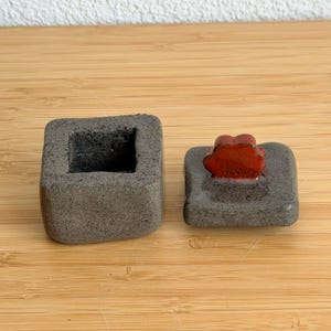 May include: A square ceramic container with a dark gray, speckled, textured finish and an open top. Beside it lies a matching square lid featuring a prominent, glossy, bright red handle shaped like a stylized flower or cloud element.