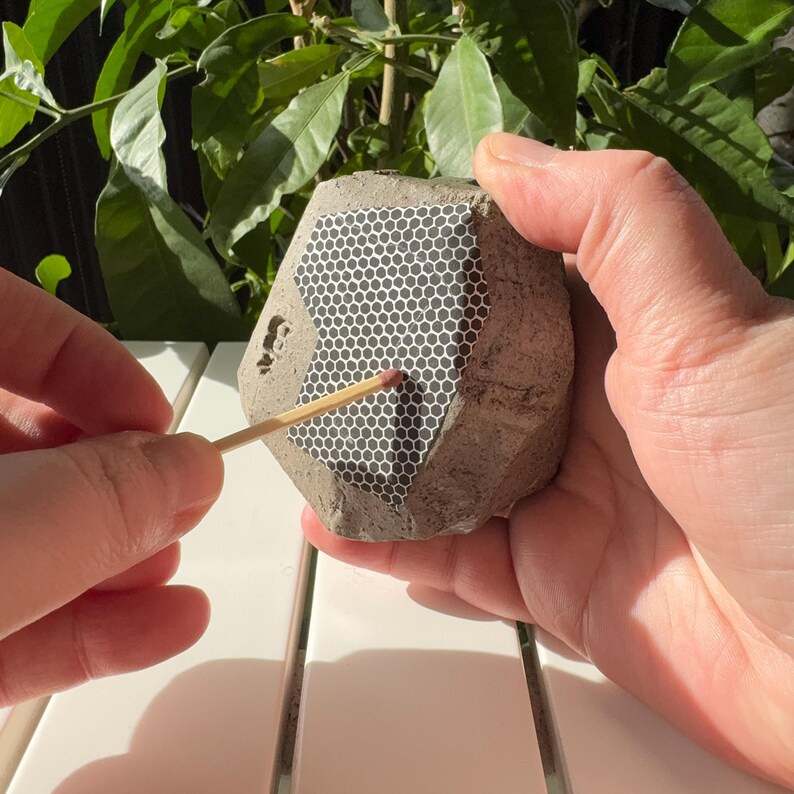 May include: A gray concrete hexagonal object with a black honeycomb pattern on one side. A wooden matchstick is being held against the honeycomb pattern.