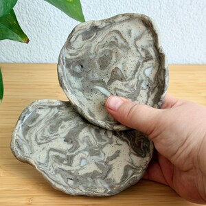 Könnte beinhalten: Zwei kleine, unregelmäßig handgeformte Keramikschalen mit einem Marmormuster aus cremefarbenen, grauen und dunkelbraunen Wirbeln. Die Oberflächen haben ein glänzendes Finish mit sichtbaren Sprenkeln im Ton und unebenen, organischen Rändern.
