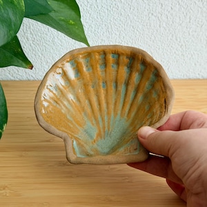 May include: A ceramic dish shaped like a scallop shell with a glossy glaze featuring a blend of warm ochre yellow and soft seafoam green colors. The surface has raised ridges radiating from the base to the scalloped edge of the shell.