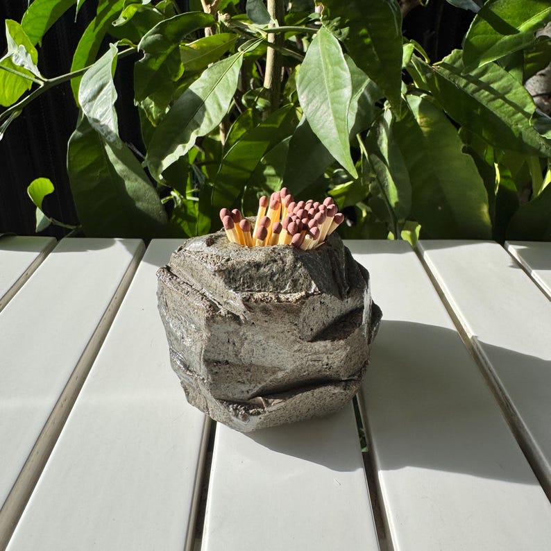 May include: A gray concrete match holder shaped like a rock with a pile of matches inside.