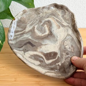 May include: A hand-formed ceramic dish with an irregular, organic shape. The surface features a marbled pattern of swirling cream, beige, and dark brown tones with subtle speckles throughout, finished with a smooth, glossy glaze.
