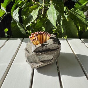 May include: A gray concrete match holder with a rough, textured surface. The holder is filled with a bundle of wooden matches with red tips.