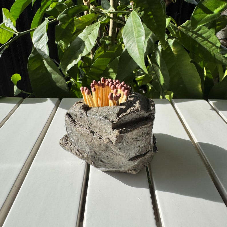 May include: A gray, rock-shaped match holder with a rough texture, filled with wooden matches with red tips. The holder is sitting on a white wooden surface.