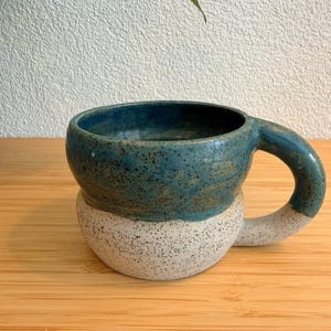 May include: Handmade ceramic mug with a rounded shape. The top half is a speckled teal, and the bottom half is speckled white. The handle is teal. The mug sits on a wooden surface.