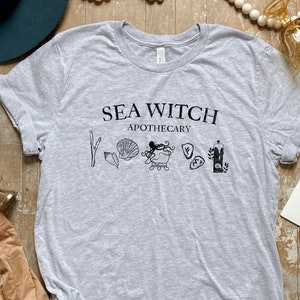 May include: Grey t-shirt with the text "Sea Witch Apothecary" and a graphic of an octopus, seashells, and other sea-themed imagery.