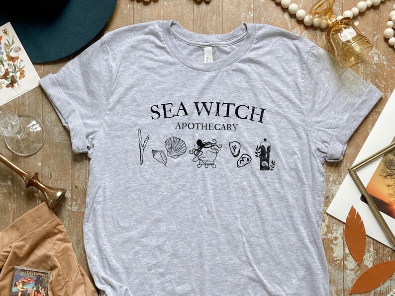 May include: Grey t-shirt with the text "Sea Witch Apothecary" and a graphic of an octopus, seashells, and other sea-themed imagery.