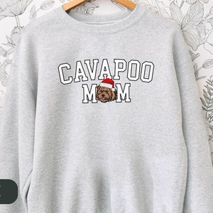 May include: A gray sweatshirt with the text "CAVAPOO MOM" in white block letters. A cartoon image of a brown cavapoo dog wearing a red Santa hat is centered above the text.