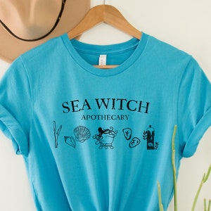 May include: A turquoise blue t-shirt with the text "Sea Witch Apothecary" in black lettering. The design includes a cauldron, seashells, seaweed, and a candle.