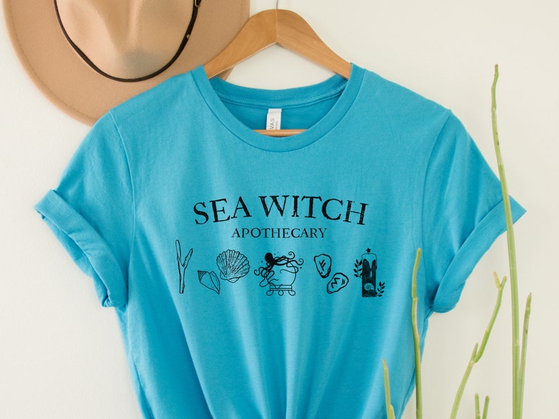 May include: A turquoise blue t-shirt with the text "Sea Witch Apothecary" in black lettering. The design includes a cauldron, seashells, seaweed, and a candle.