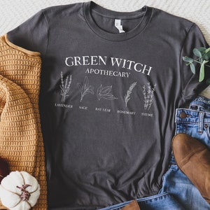 May include: A dark gray t-shirt with the text "Green Witch Apothecary" and illustrations of herbs, including lavender, sage, bay leaf, rosemary, and thyme.