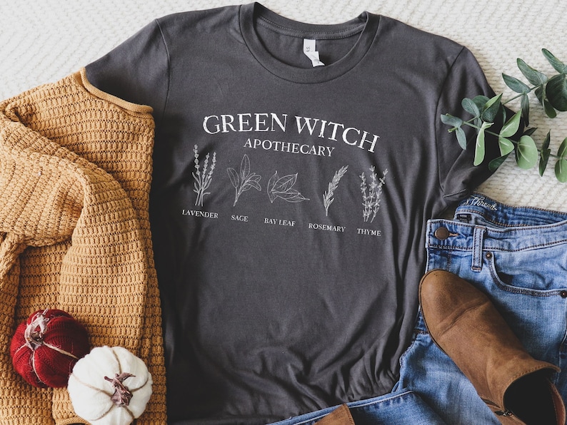May include: A dark gray t-shirt with the text "Green Witch Apothecary" and illustrations of herbs, including lavender, sage, bay leaf, rosemary, and thyme.