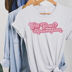 May include: White t-shirt with pink text that reads "Get Your Mammogram" in a retro font.