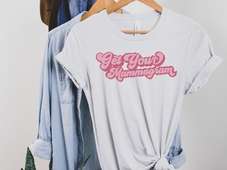 May include: White t-shirt with pink text that reads "Get Your Mammogram" in a retro font.