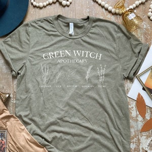May include: Green Witch Apothecary t-shirt in a muted green color with white text and illustrations of herbs. The text reads "Green Witch Apothecary" with the names of herbs listed below: Lavender, Sage, Bay Leaf, Rosemary, Thyme.