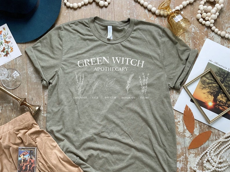 May include: Green Witch Apothecary t-shirt in a muted green color with white text and illustrations of herbs. The text reads "Green Witch Apothecary" with the names of herbs listed below: Lavender, Sage, Bay Leaf, Rosemary, Thyme.
