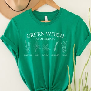 May include: Green t-shirt with the text "Green Witch Apothecary" and illustrations of herbs labeled "Lavender", "Sage", "Bay Leaf", "Rosemary", and "Thyme".