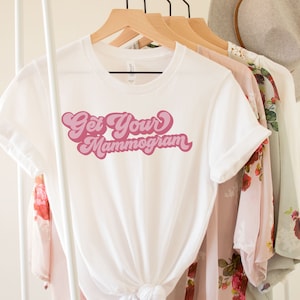 May include: White t-shirt with pink text that reads "Get Your Mammogram" in a retro font.