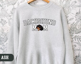 May include: A gray sweatshirt with the text "Dachshund Mom" in white letters and a black and brown dachshund illustration. The sweatshirt is available in the color ash.