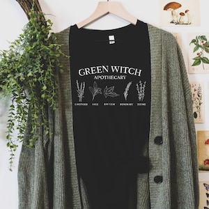 May include: Black t-shirt with white text that reads "Green Witch Apothecary" and illustrations of herbs labeled "Lavender", "Sage", "Bay Leaf", "Rosemary", and "Thyme".