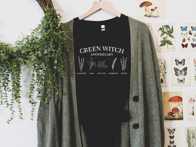 May include: Black t-shirt with white text that reads "Green Witch Apothecary" and illustrations of herbs labeled "Lavender", "Sage", "Bay Leaf", "Rosemary", and "Thyme".