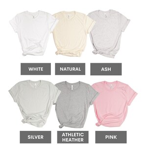 May include: Six different color t-shirts, including white, natural, ash, silver, athletic heather, and pink. The shirts are all folded and laid out on a white background.