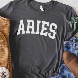 K&ouml;nnte beinhalten: Ein dunkelgraues T-Shirt mit dem Wort "ARIES" in wei&szlig; auf der Vorderseite. Das Shirt liegt flach auf einer wei&szlig;en Oberfl&auml;che mit einem braunen Pullover, einem wei&szlig;en K&uuml;rbis und einer blauen Jeans.