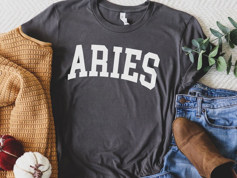 K&ouml;nnte beinhalten: Ein dunkelgraues T-Shirt mit dem Wort "ARIES" in wei&szlig; auf der Vorderseite. Das Shirt liegt flach auf einer wei&szlig;en Oberfl&auml;che mit einem braunen Pullover, einem wei&szlig;en K&uuml;rbis und einer blauen Jeans.