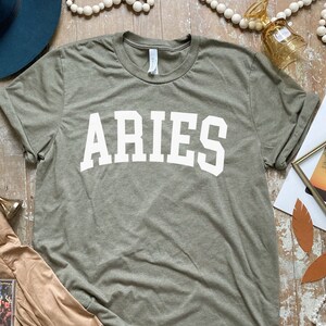 K&ouml;nnte beinhalten: Ein gr&uuml;nes T-Shirt mit dem Wort "ARIES" in wei&szlig; auf der Vorderseite.