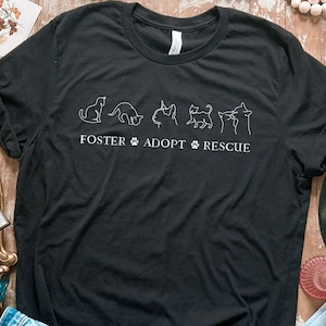 May include: Black t-shirt with a white outline of five cats and the text "FOSTER ADOPT RESCUE" with a paw print between each word.